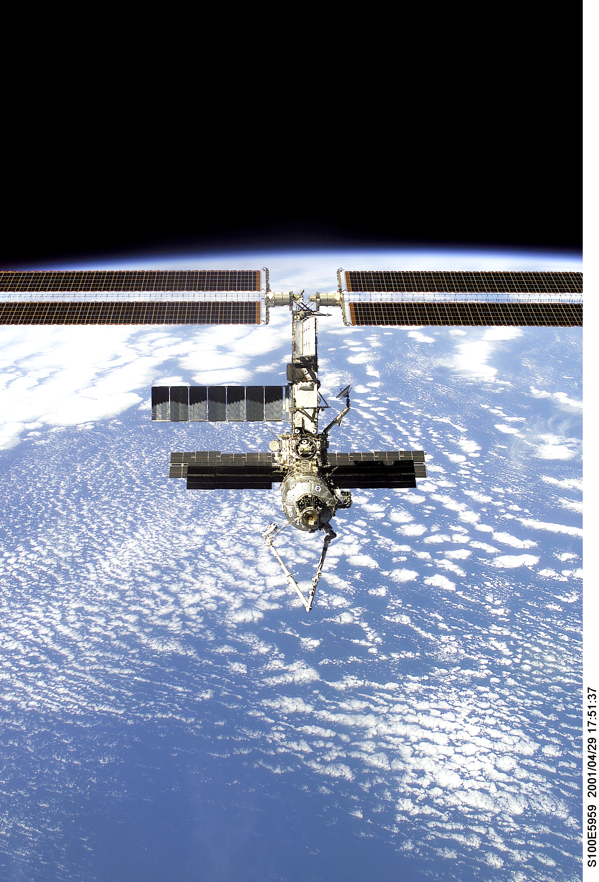 The International Space Station photographed from Space Shuttle Endeavour during undocking, April 2001. Solar arrays extend outward against Earth&rsquo;s cloud-covered surface below.